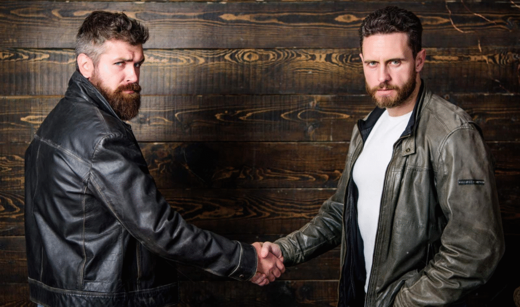 Two men wearing rugged leather jackets shaking hands confidently, set against a rustic wooden background